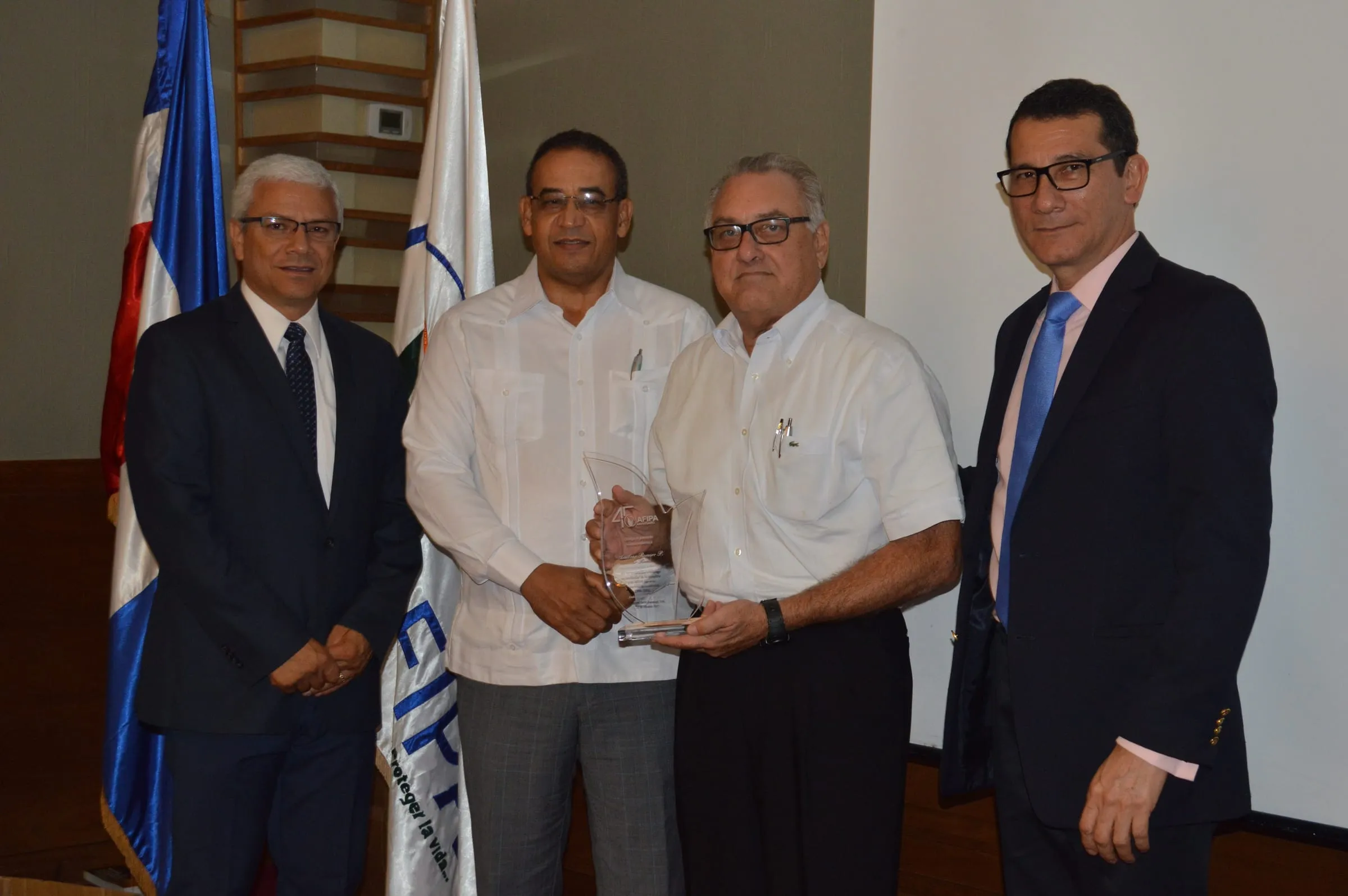 Dos expresidentes de AFIPA reciben su placa de reconocimiento acompañados por directivos, delante de la bandera dominicana