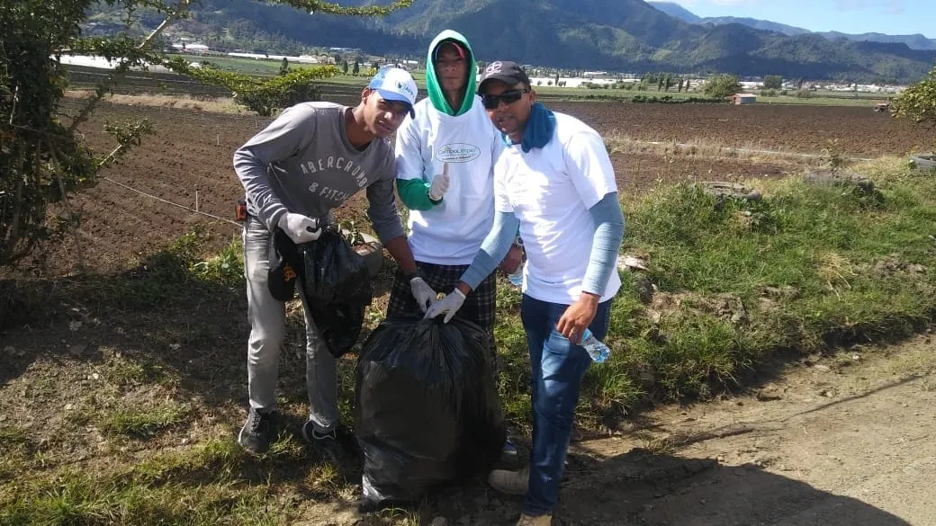 Tres voluntarios con guantes llenan una funda de envases recogidos junto a un campo recién arado de Tireo, con las montañas de Constanza detrás