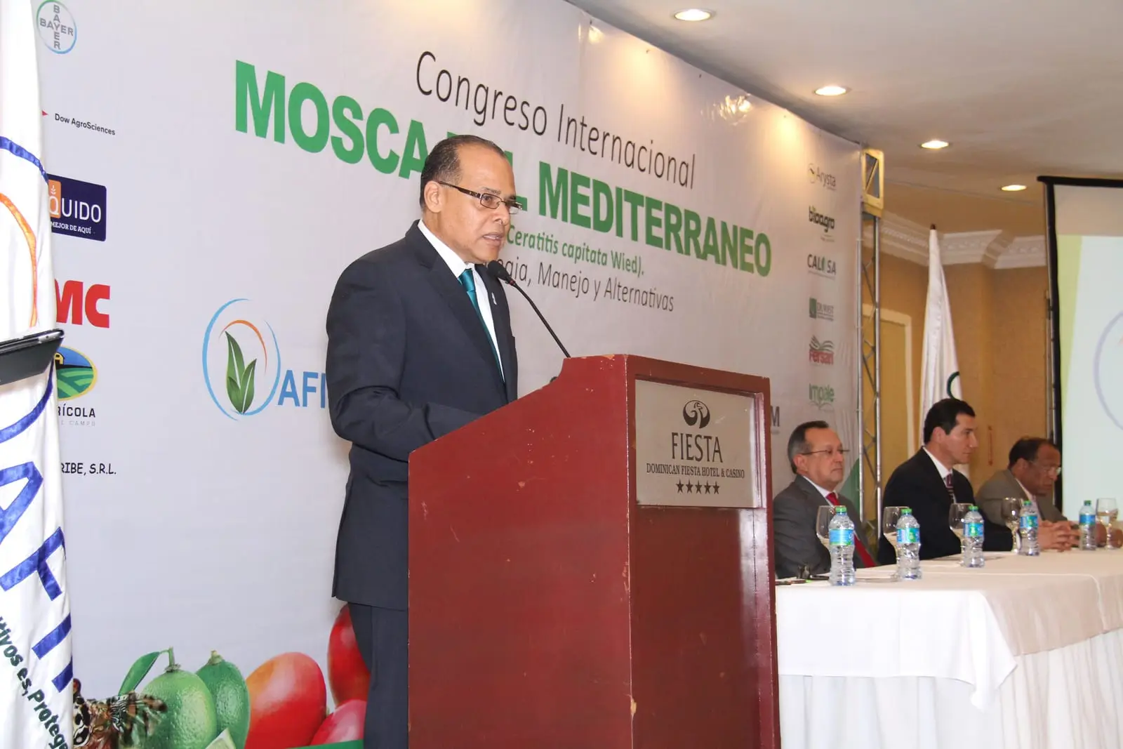 Un expositor habla desde el pódium bajo el banner del Congreso Internacional Mosca del Mediterráneo, con la mesa de honor sentada a su lado