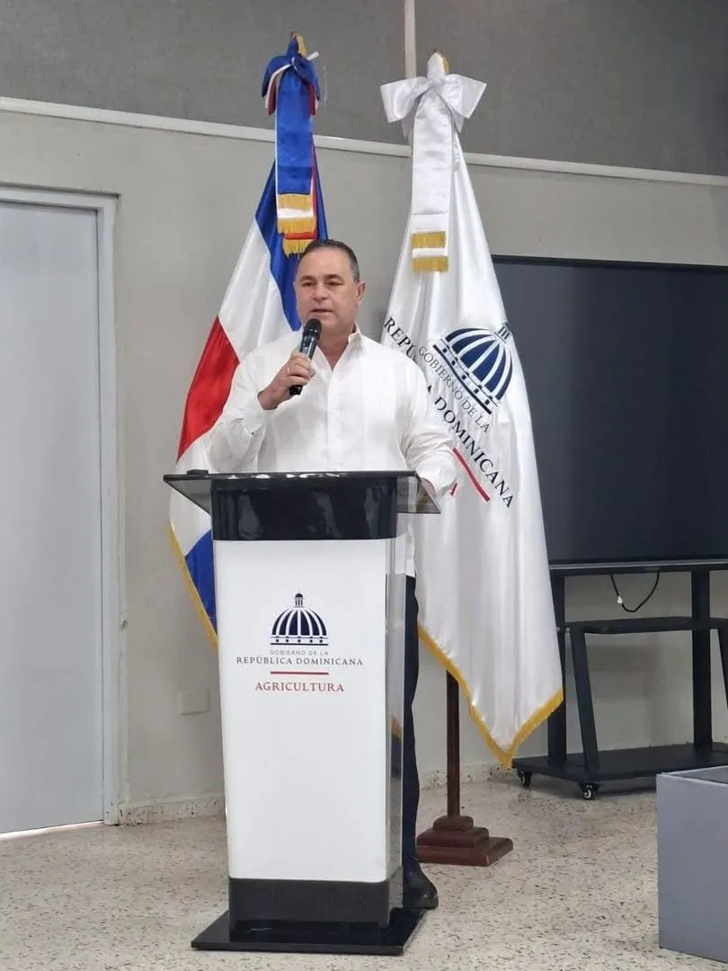 El ministro de Agricultura habla desde el podio, entre la bandera dominicana y la del gobierno